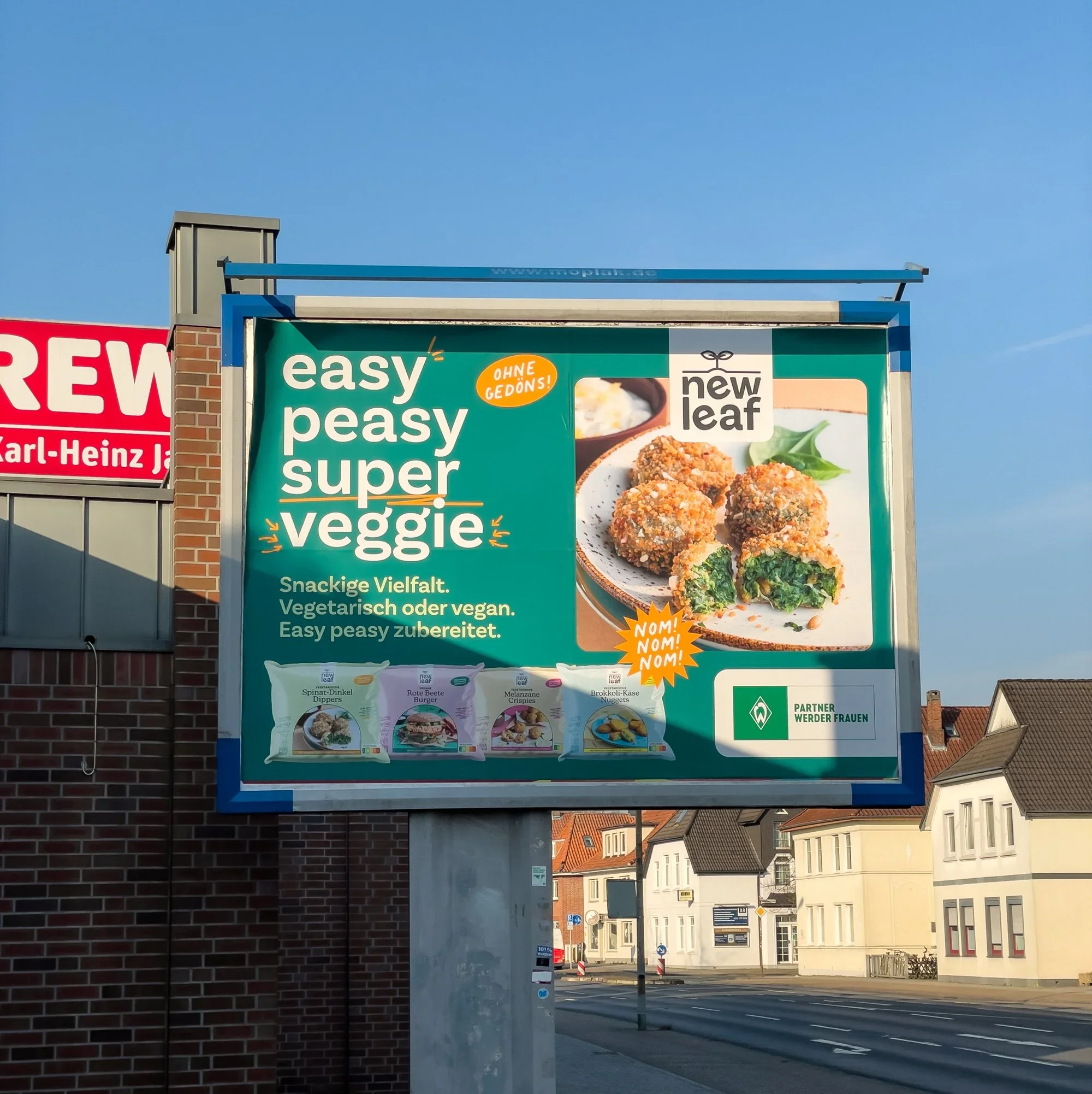Straßenbahnhaltestelle mit einem großen, grünen Werbeschild für vegetarisches, veganes Essen, das Falafel und andere Gerichte zeigt, in einer Stadtumgebung mit Wohnhäusern.