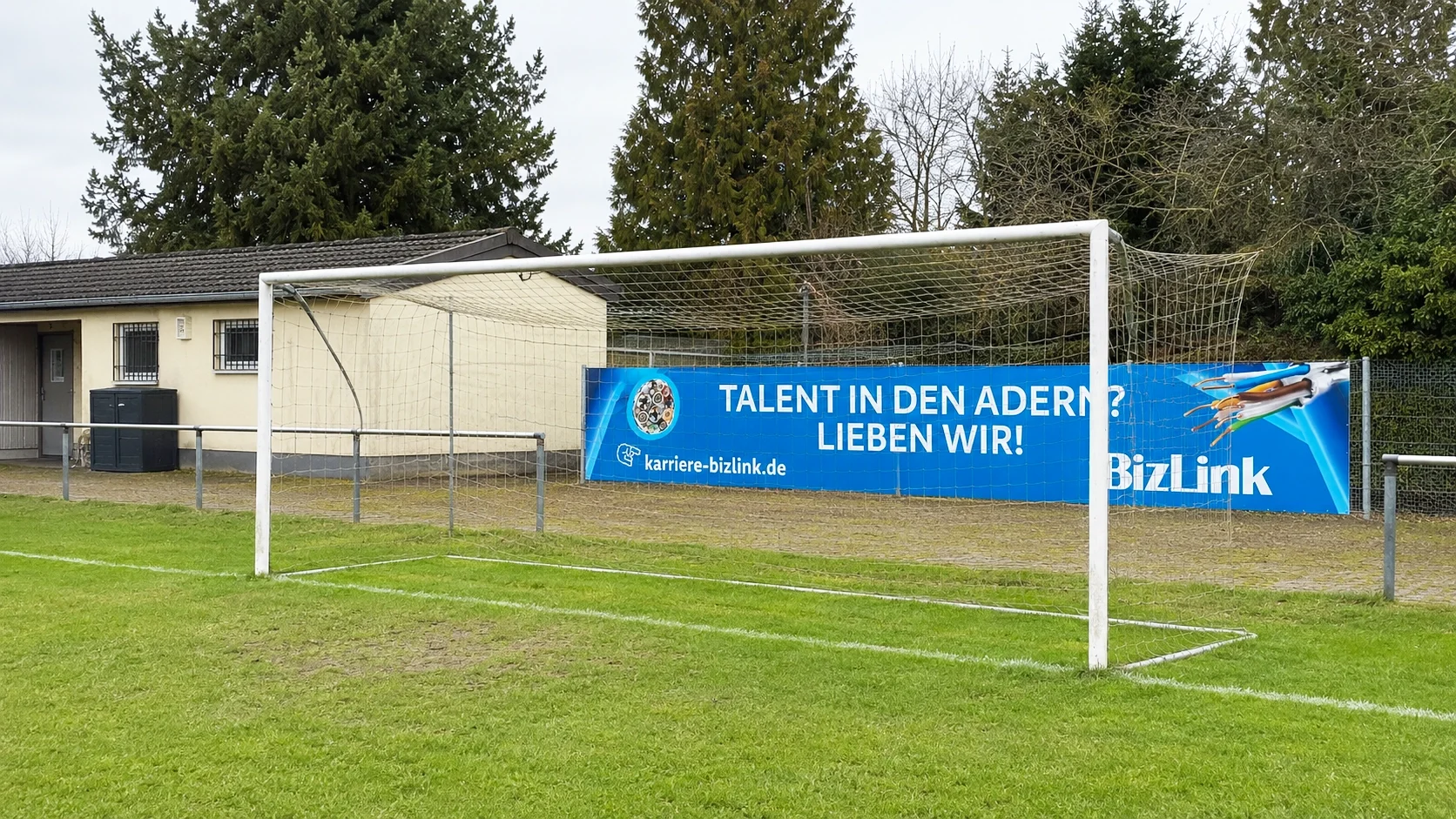 Werbetafel vor einem Fußballtor mit einer blauen Werbung, die einen Computerchip und Kabel zeigt, mit dem Text 'Talent in den Adern? Lieben wir!'.