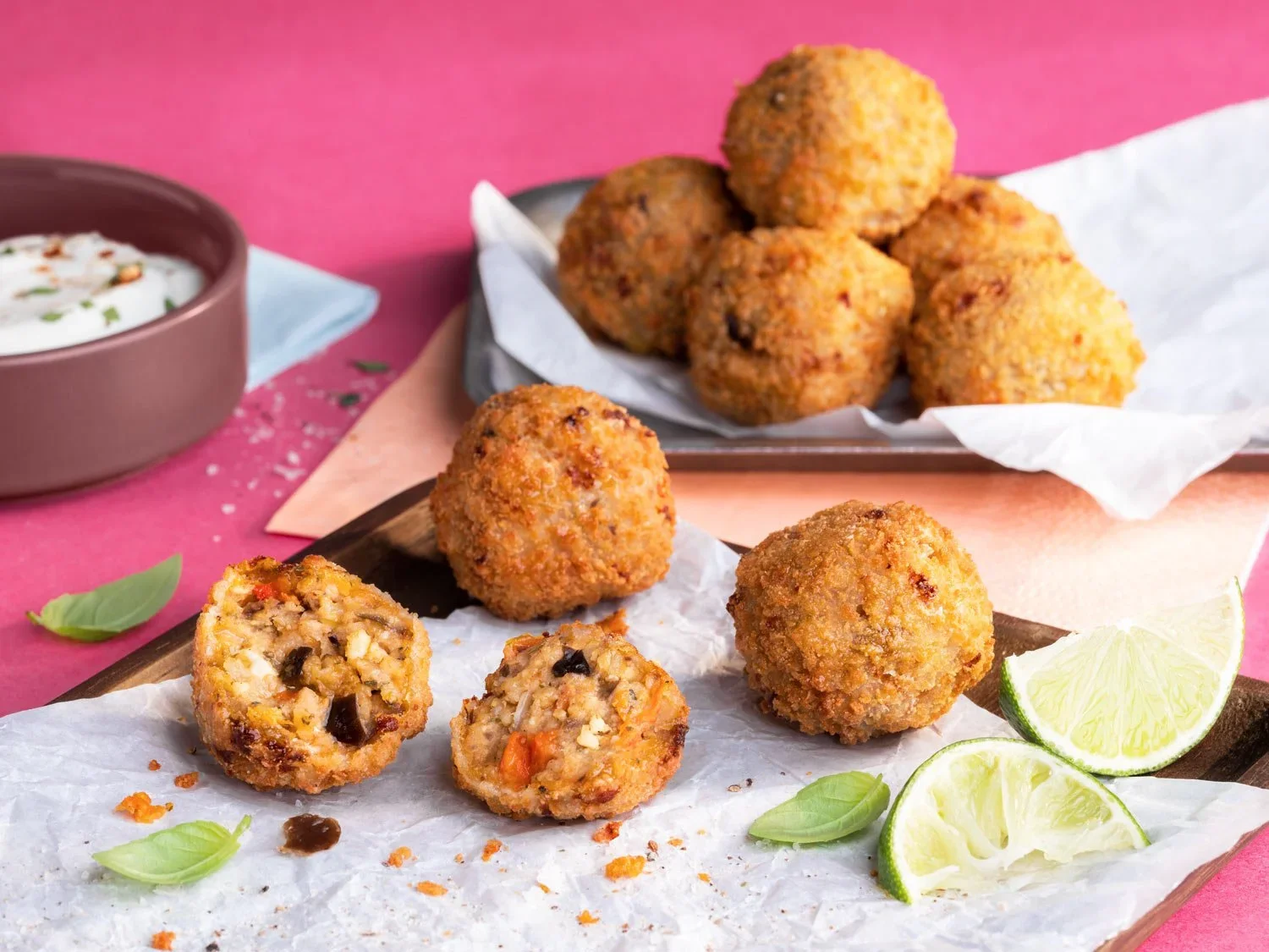 Krustenartige Knödel, gefüllt mit Hackfleisch, serviert mit Limettenscheiben und frischen Basilikumblättern, auf weißem Papier auf einem Holzbrett, mit einer Schüssel Joghurt-Dip im Hintergrund auf einem pinken Tisch.