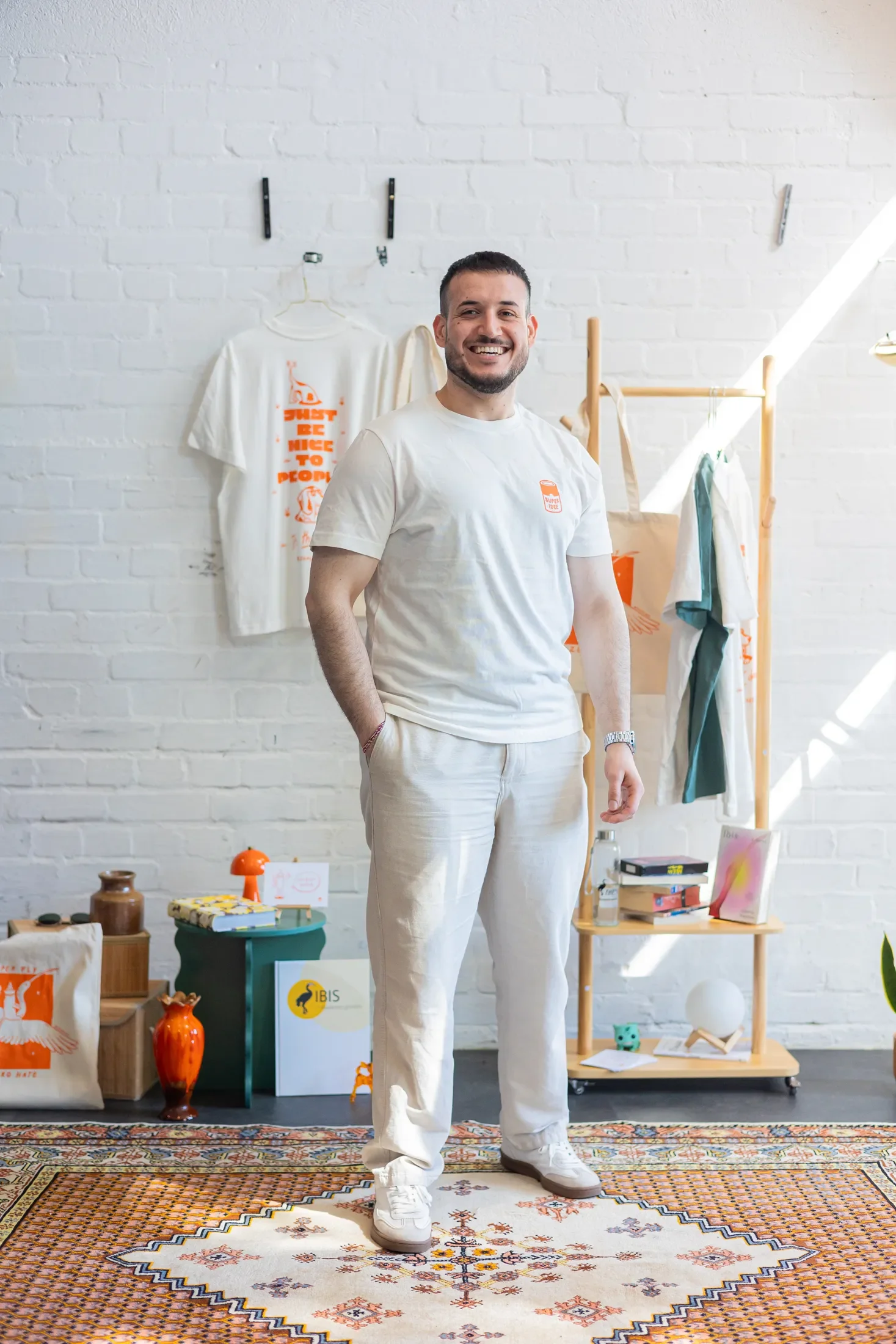 Ein Mann in weißem T-Shirt lächelt in die Kamera. Hinter ihm hängt an einem Bügel ein T-Shirt mit der Aufschrift "Just be nice to People"