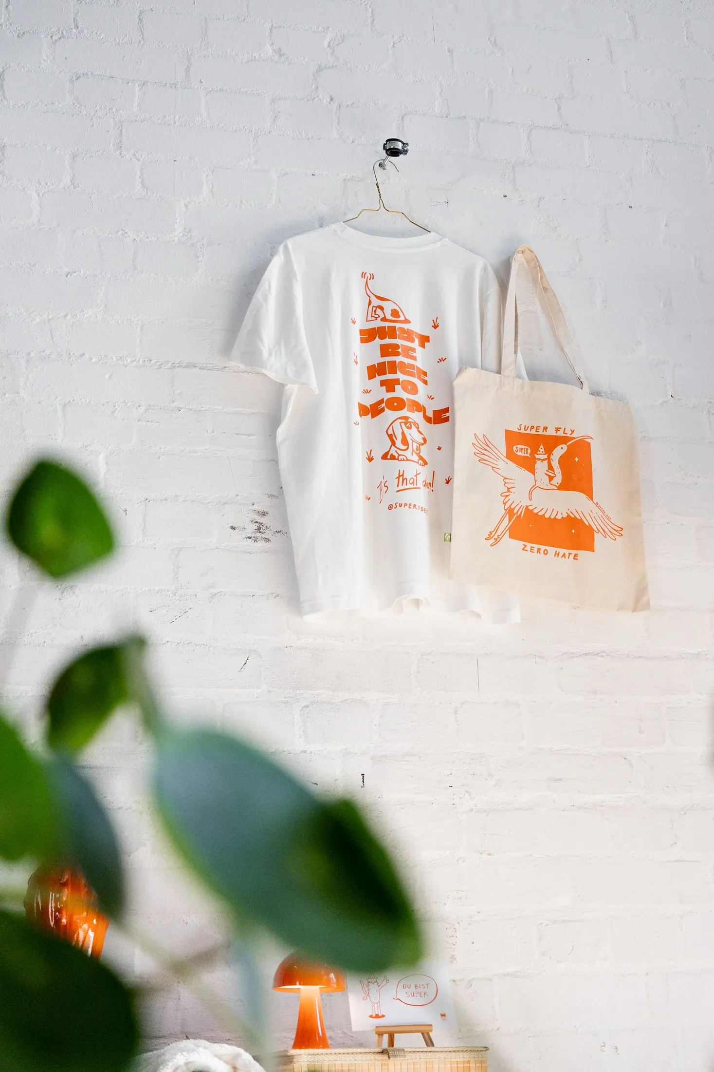 Weiße Wand mit einem weißen T-Shirt mit orangenem Druck, einer Tasche mit orangefarbenem Druck und einem kleinen weißen Poster mit rotem Schriftzug im Hintergrund. Im Vordergrund sind unscharfe grüne Blätter.