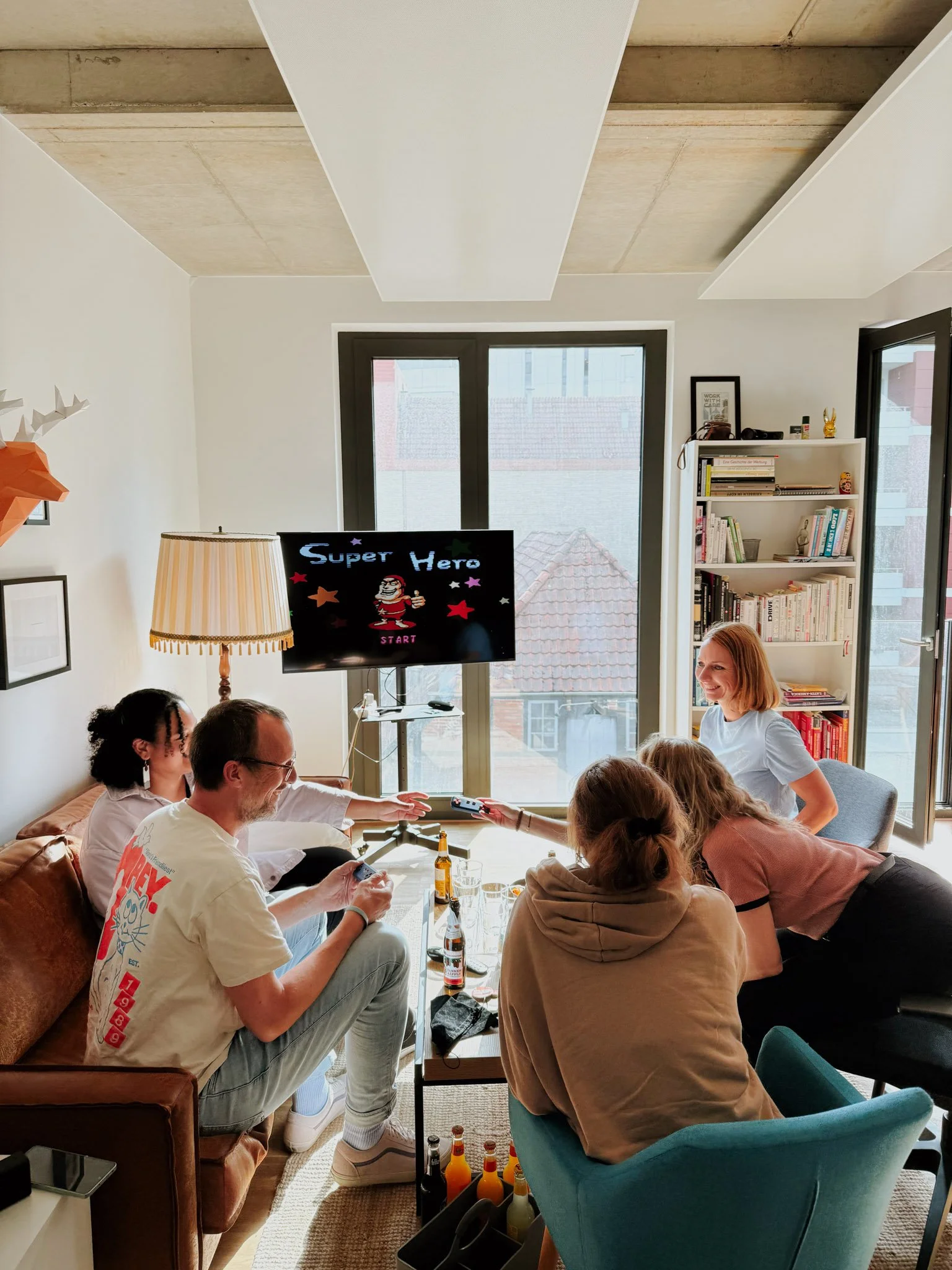Mehrere Menschen sitzen in einem Wohnzimmer, spielen ein Videospiel namens "Super Hero" und unterhalten sich. Im Hintergrund große Fenster, eine weiße Bücherregal und eine Stehlampe.