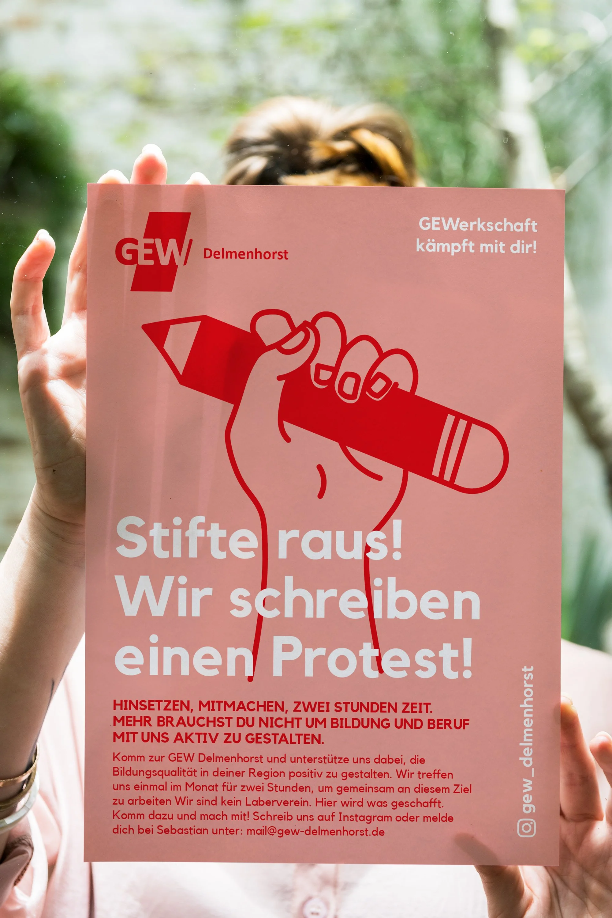 Ein handgehaltenes Poster mit der Aufschrift 'Stifte raus! Wir schreiben einen Protest!' und einem roten Projektionszeichen, das eine Hand hält, die einen Stift fasst. Es gibt einen Textblock mit weiteren Informationen und Kontaktdaten auf der unteren Hälfte des Posters.