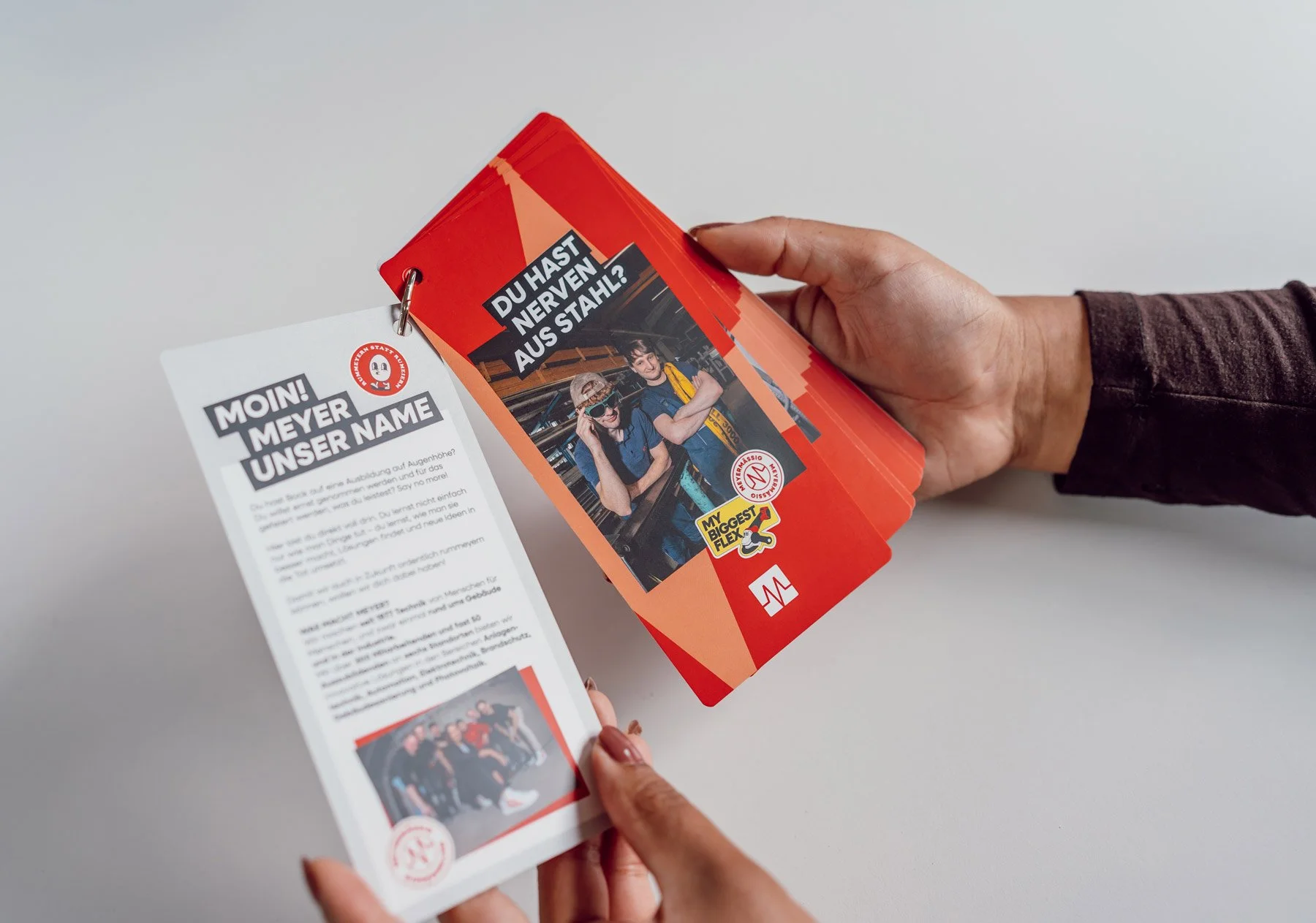 Eine Hand hält eine rote Broschüre mit einem Bild von zwei jungen Menschen in Schutzkleidung und Überschrift 'Du hast Nerven aus Stahl?'. Im Hintergrund liegt eine weitere moree Broschüre auf einem weißen Tisch.