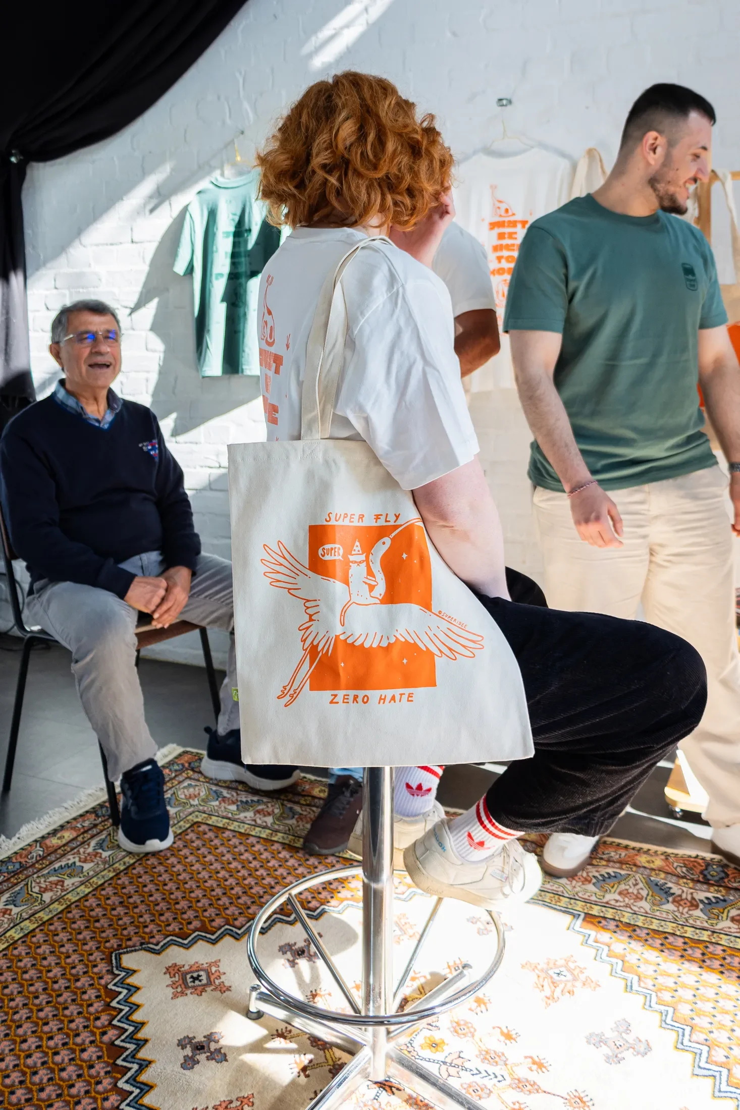 Person sitzt entspannt auf einem Hocker, mit der Super-Tasche unterm Arm und unterhält sich mit drei weiteren Personen