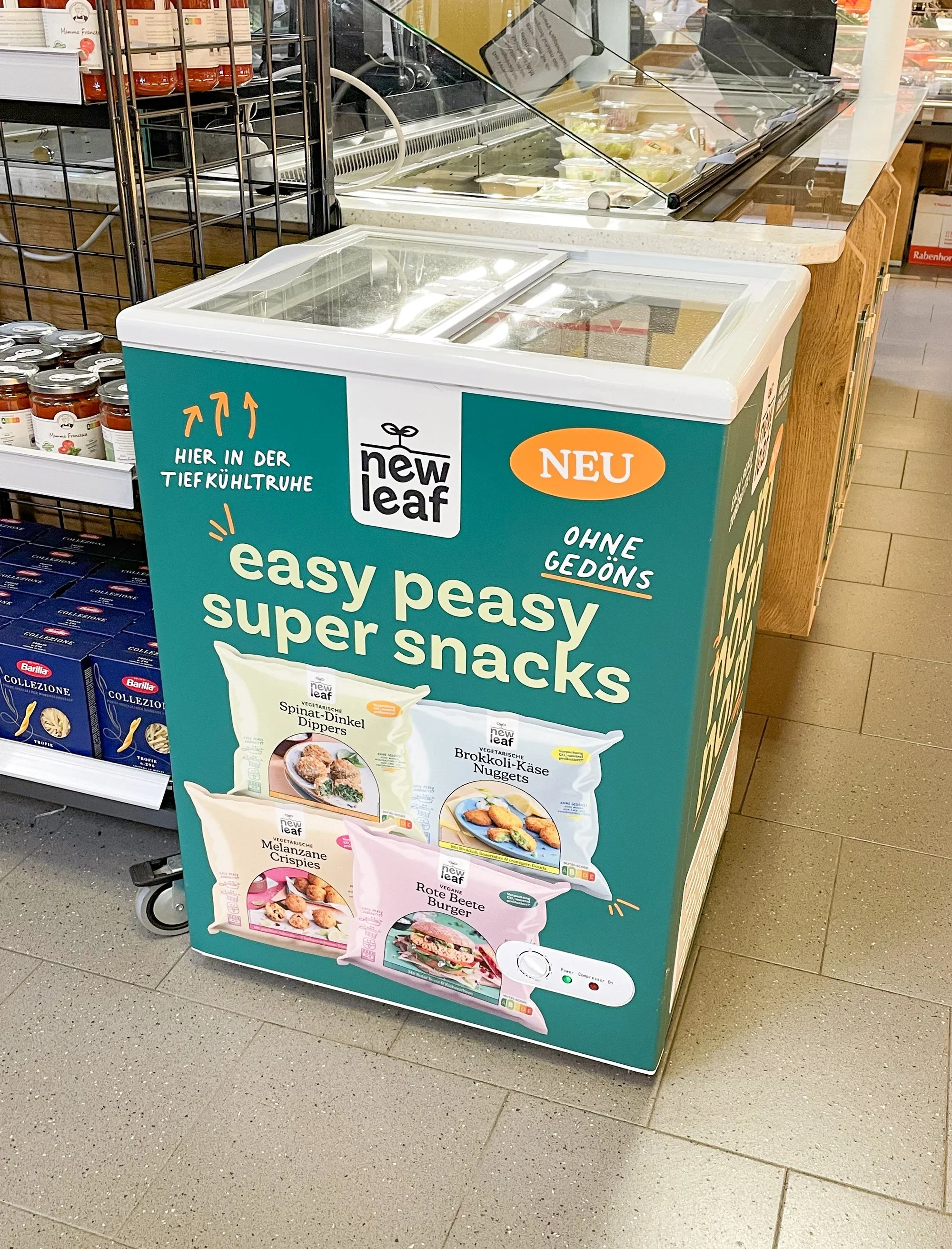 Ein Kühlgerät im Supermarkt mit verschiedenen Vorräten und einem Aufsteller, der vegane Tiefkühlprodukte von der Marke 'new leaf' bewirbt.
