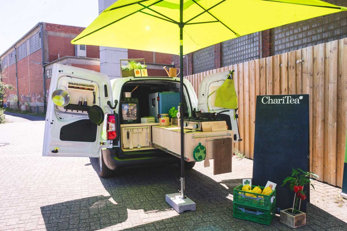 Das Mampfmobil – Ein mobiler Küchenstand im Freien mit einem großen gelben Sonnenschirm, einer Theke, einem Kühlschrank, Topf, Pflanzen und einem Chalkboard.
