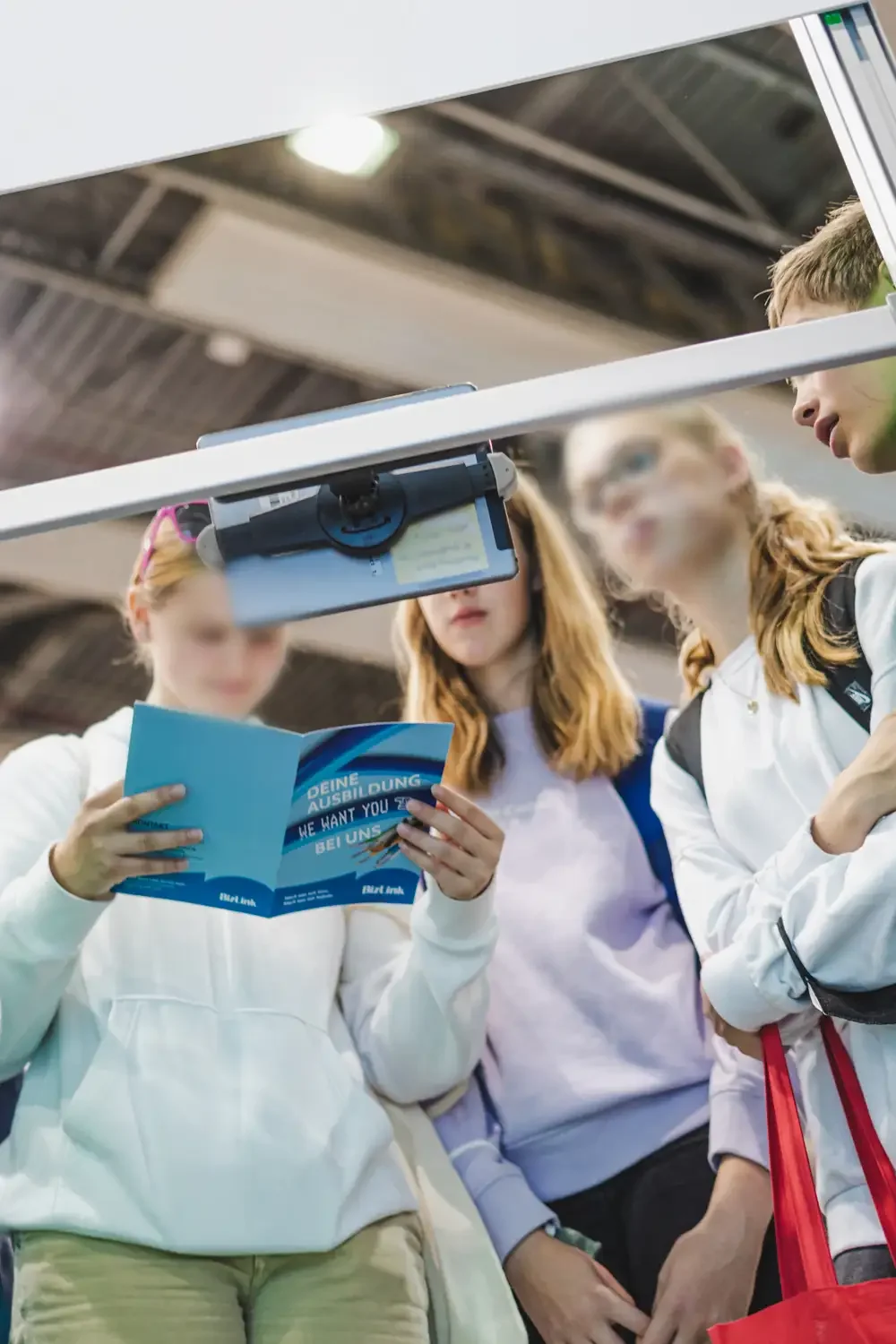 Vier junge Menschen stehen im Gespräch und schauen auf eine Broschüre, während ein Smartphone an eine Halterung über ihnen befestigt ist.