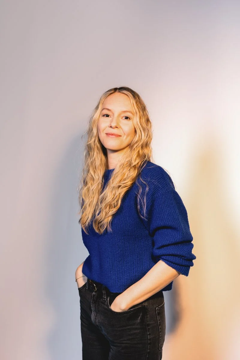 Junge Frau mit blonden, lockigen Haaren trägt eine blaue Sweater und schwarze Jeans, lächelt vor einer neutralen Wand.