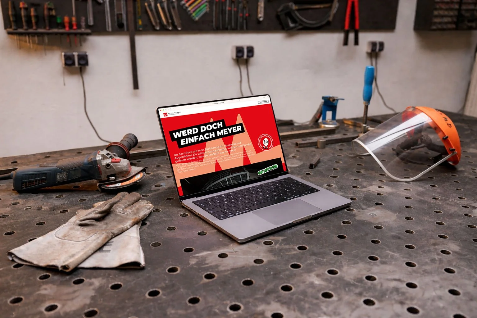 Werkbank in einer Werkstatt mit einem Laptop, Arbeitshandschuhen, einem Schleifgerät, einem Kopfschutz, einem Schraubenzieher und anderen Werkzeugen.
