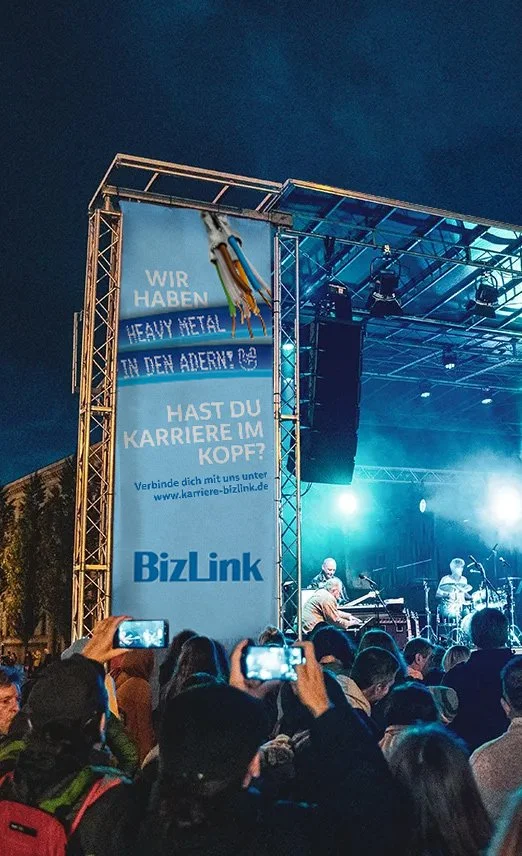 Menschen vor einer Bühne bei einem Live-Konzert mit einer großen Werbetafel. Die Tafel trägt den Text: "Wir haben Heavy Metal in den Adern! Hast du Karriere im Kopf?" und das BizLink-Logo.