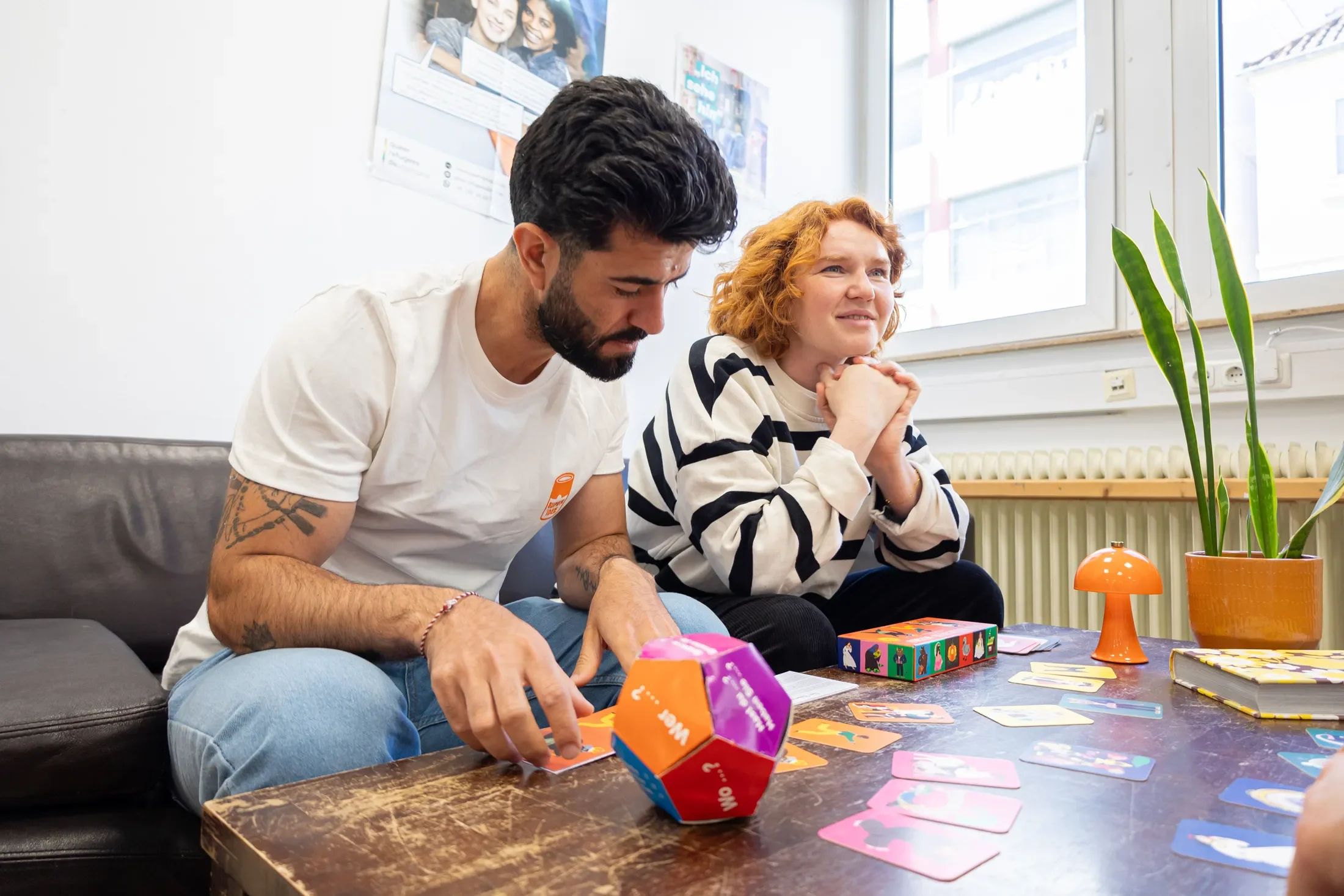 Zwei Menschen spielen ein Gesellschaftsspiel an einem Esstisch. Der Mann hat dunkle Haare, einen Bart und trägt ein weißes T-Shirt, das eine Orange Note hat. Die Frau hat rote Locken und trägt ein schwarz-weiß gestreiftes Hemd. Es gibt bunte Spielkarten, Würfel und dekorative Gegenstände auf dem Tisch, während im Hintergrund ein Fenster und eine Pinnwand zu sehen sind.