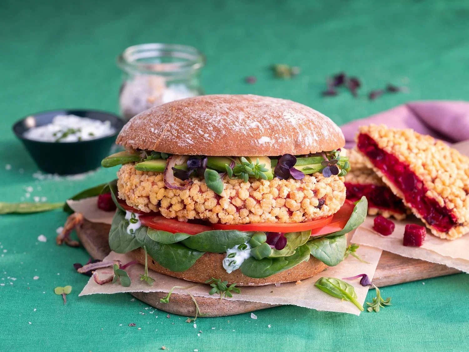 Ein Sandwich mit frittiertem Hähnchen, Tomate, Salat und Sprossen auf einem Holzteller, im Hintergrund eine Limonade und ein Stück Kirschkuchen.