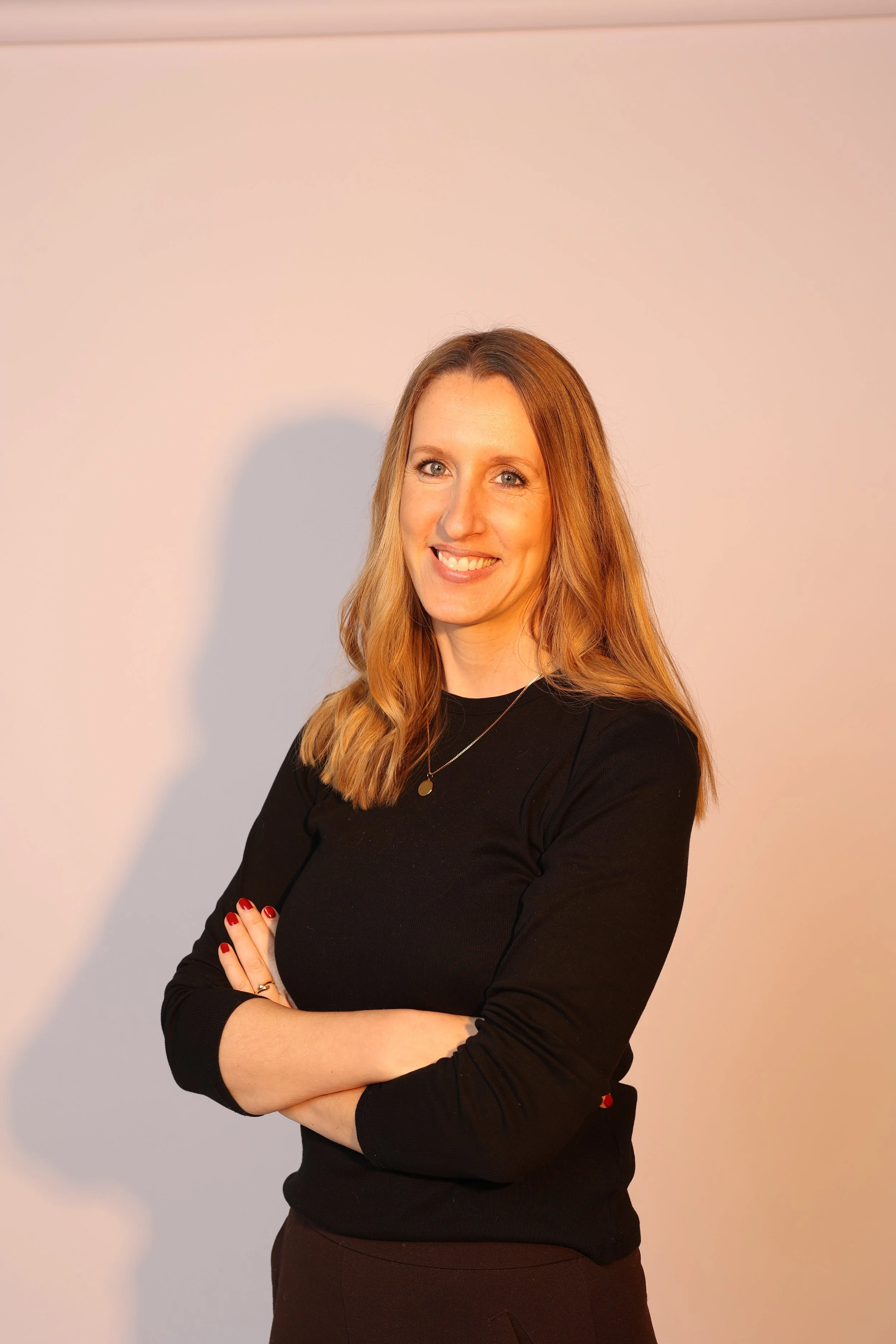 Junge Frau mit langem, gewelltem Haar und einem schwarzen Pullover, lächelnd, Arme verschränkt, vor hellem, neutralem Hintergrund.