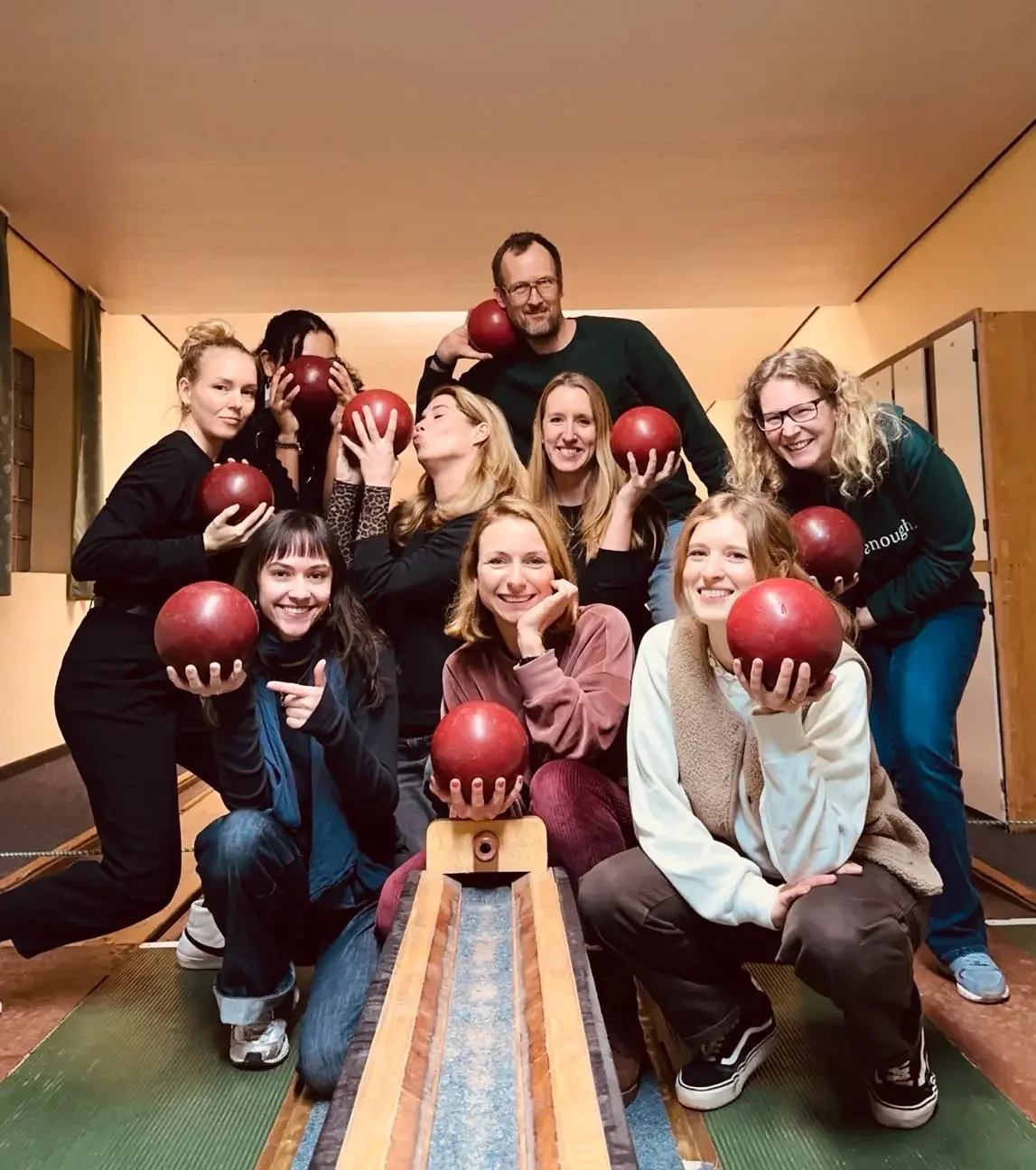 Gruppenfoto von zehn Menschen, die in einem Raum mit Bowlingbahn sitzen und mit roten Bowlingkugeln posieren, lachen und Spaß haben.