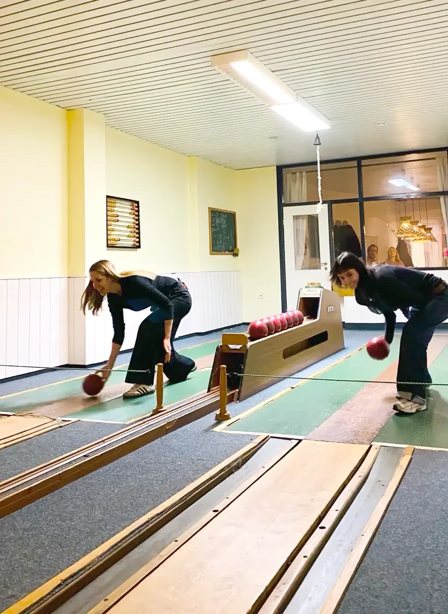 Zwei Frauen spielen Bowling in einer Lichtdurchfluteten Bowlingbahn, während im Hintergrund einige Leute durch ein Fenster zuschauen.