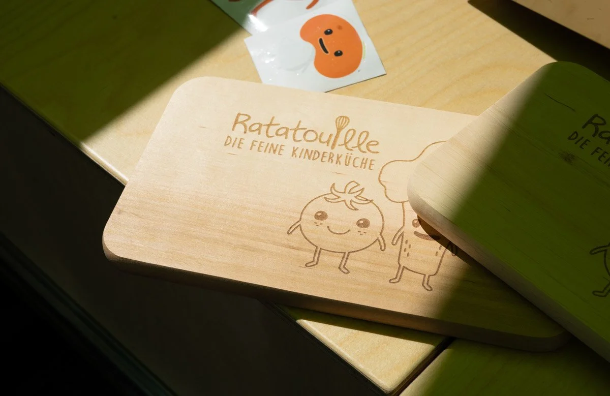 Holzbrett mit dem Text 'Ratatouille – Die feine Kinderküche' und einer niedlichen Zeichnung eines zufriedenen Gemüses und eines Kochs, Schattierung durch Sonnenlicht.