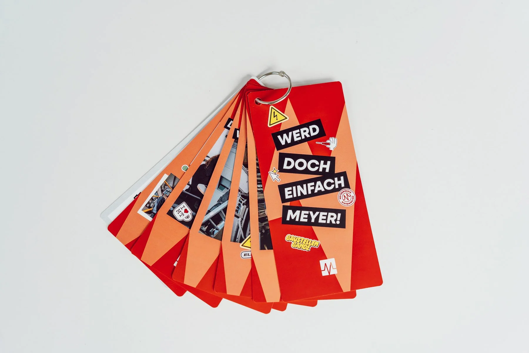 Eine Farbkarte mit roten und orangefarbenen Karten, die verschiedene Sticker und Fotos enthält, mit dem Text 'WERD DOCH EINFACH MEYER!' auf der Vorderseite.
