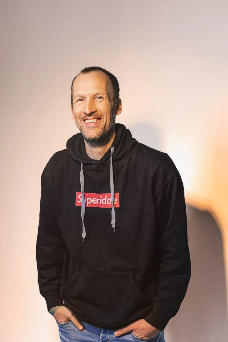Ein lächelnder Mann mit kurzem Bart, schwarzen Haaren und blauen Augen, trägt einen schwarzen Hoodie mit 'Superdry' Logo und blaue Jeans, steht vor einem neutralen Hintergrund.