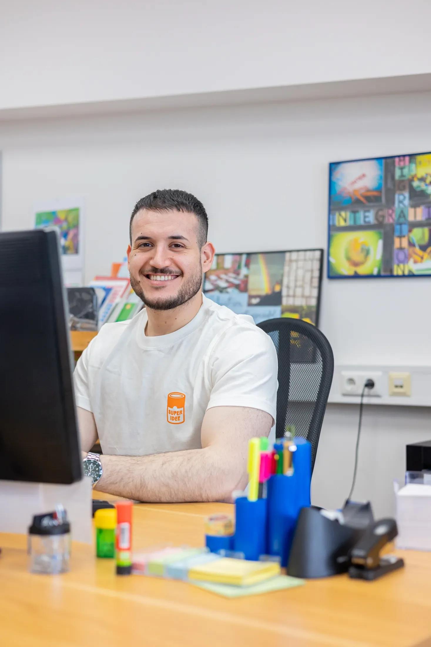 Junge Mann arbeitet in einem modernen Büro an einem Computer, mit bunten Büroartikeln und bunten Post-its auf dem Schreibtisch, hinter ihm bunte Bilder an der Wand.