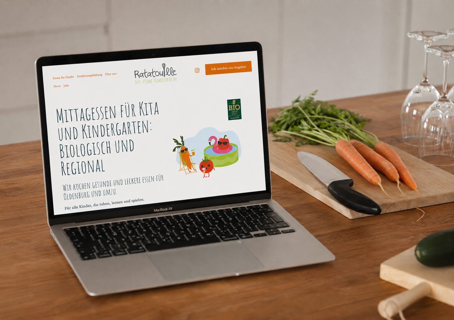 Ein Laptop zeigt eine Webseite mit dem Titel: Mittagessen für Kita und Kindergarten: Biologisch und Regional. Auf der Webseite sind bunte Skizzen von Gemüse und Wasserspielzeug. Der Tisch hat Karotten, eine Gurke, ein Messer und Weingläser.
