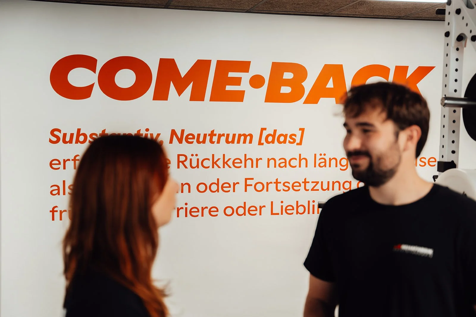 Zwei Menschen sprechen vor einem großen weißen Poster mit orange-rote Beschriftung, das das Wort 'COME-BACK' und eine Definition des Begriffs enthält.