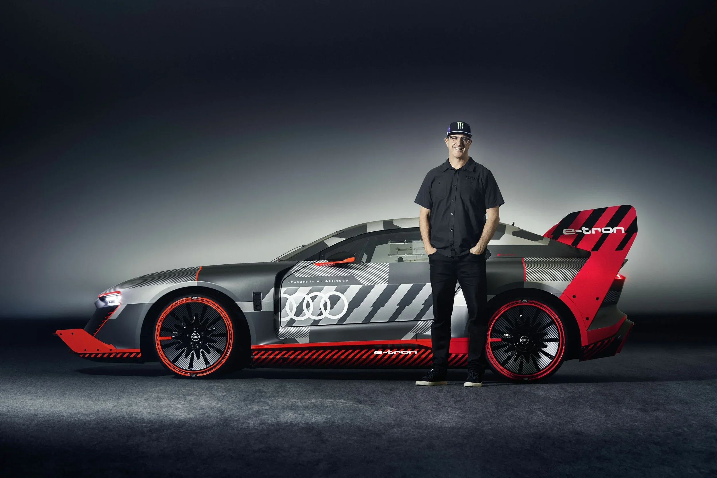 Auf dem Bild ist ein Audi S1 e-tron quattro Hoonitron Concept Car in Grau mit roten und schwarzen Akzenten zu sehen. Ein Mann in schwarzem Outfit und Cap steht lächelnd seitlich neben dem Fahrzeug vor dunklem Studiohintergrund.