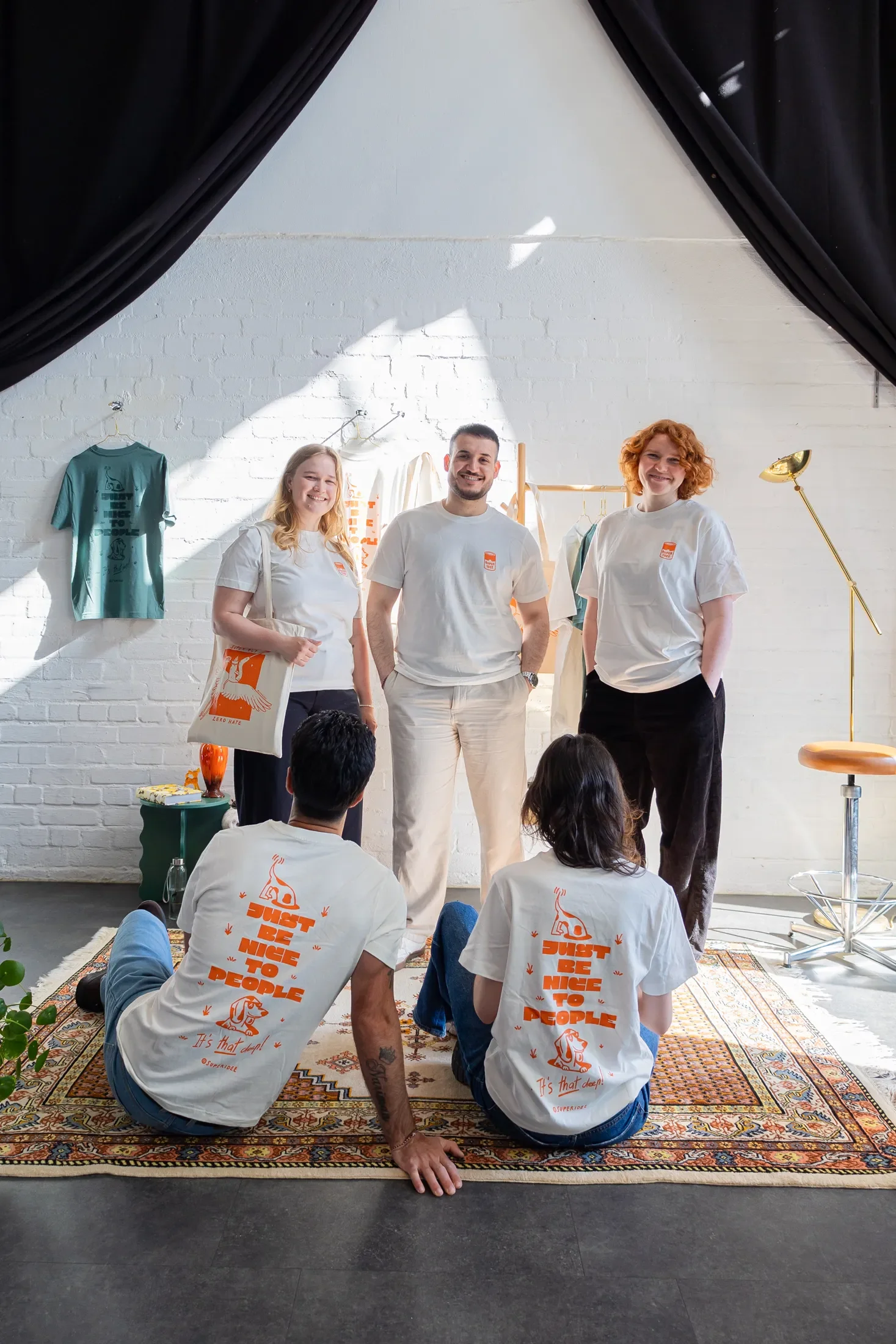Drei Menschen stehen vorne im Raum, während sechs Personen auf einer Teppichmatte sitzen. Alle tragen weiße T-Shirts mit einem orangefarbenen Logo und dem Schriftzug 'JUST BE NICE TO PEOPLE'. Die Gruppe befindet sich in einem hell erleuchteten Raum mit weißen Ziegelwänden, davor hängen T-Shirts und es gibt einen Stehlampen. Das Bild zeigt eine freundliche, informelle Zusammenkunft.
