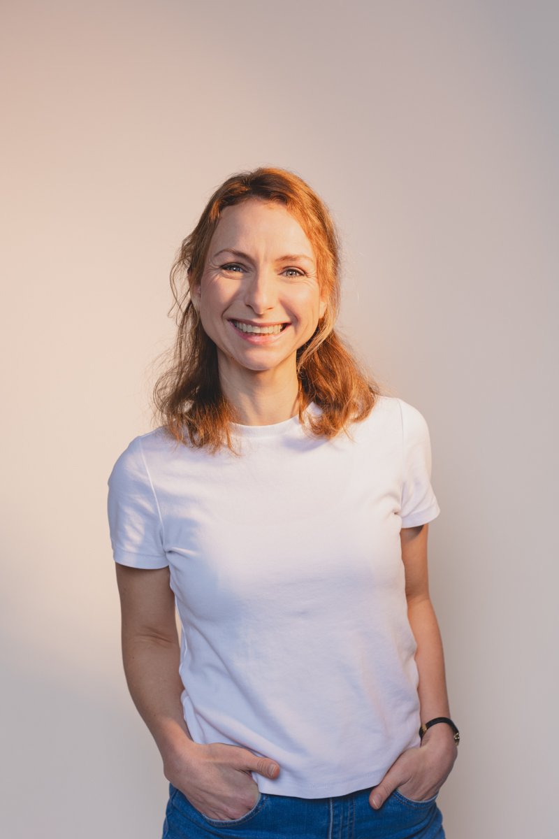 Lächelnde junge Frau mit rotbraunen Haaren, weißem T-Shirt und blauen Jeans vor neutralem Hintergrund.