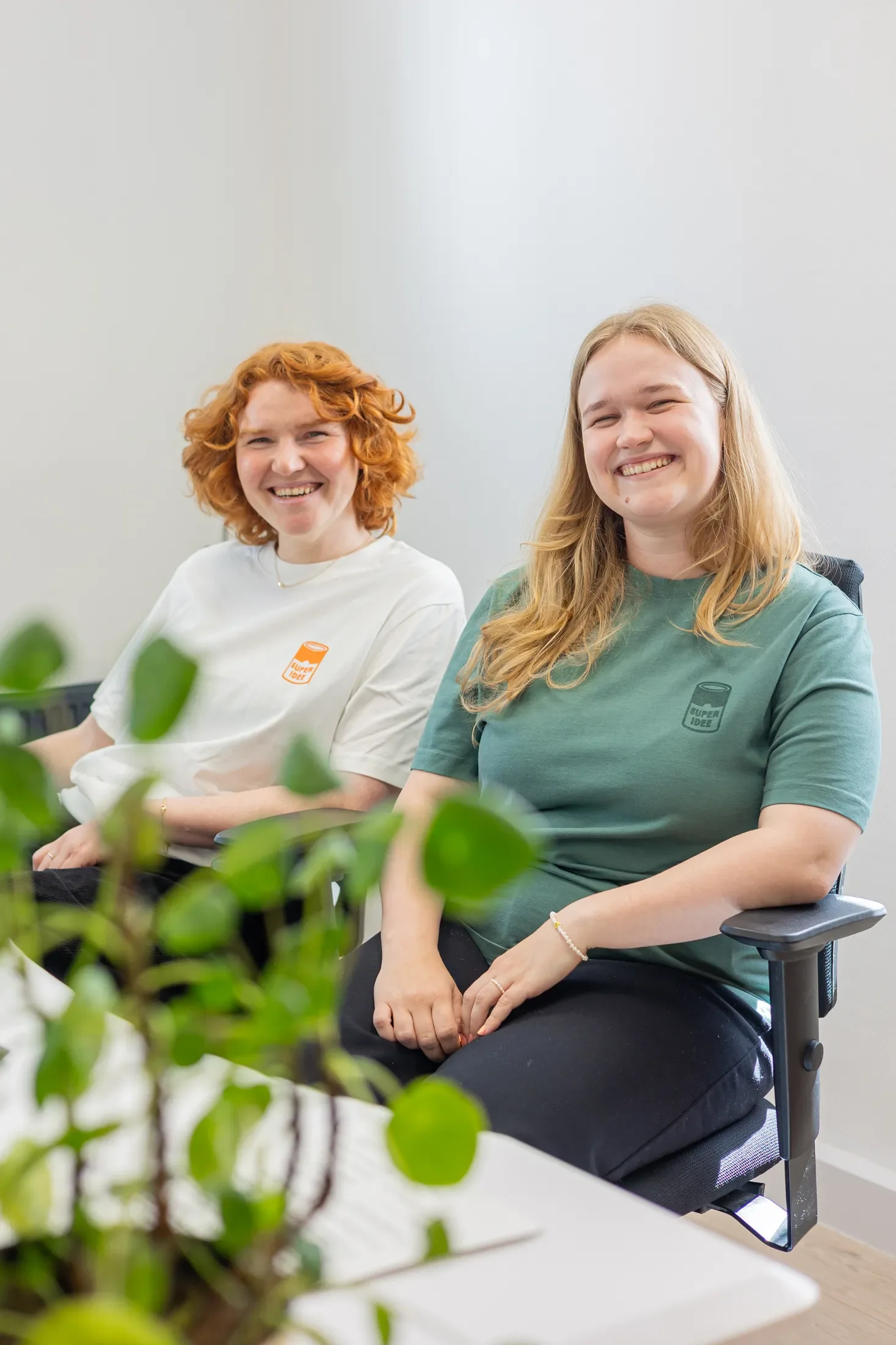 Zwei lachende Frauen sitzen in einem modernen Büro, eine trägt ein grünes T-Shirt, die andere ein weißes T-Shirt, im Vordergrund sind grüne Pflanzen.