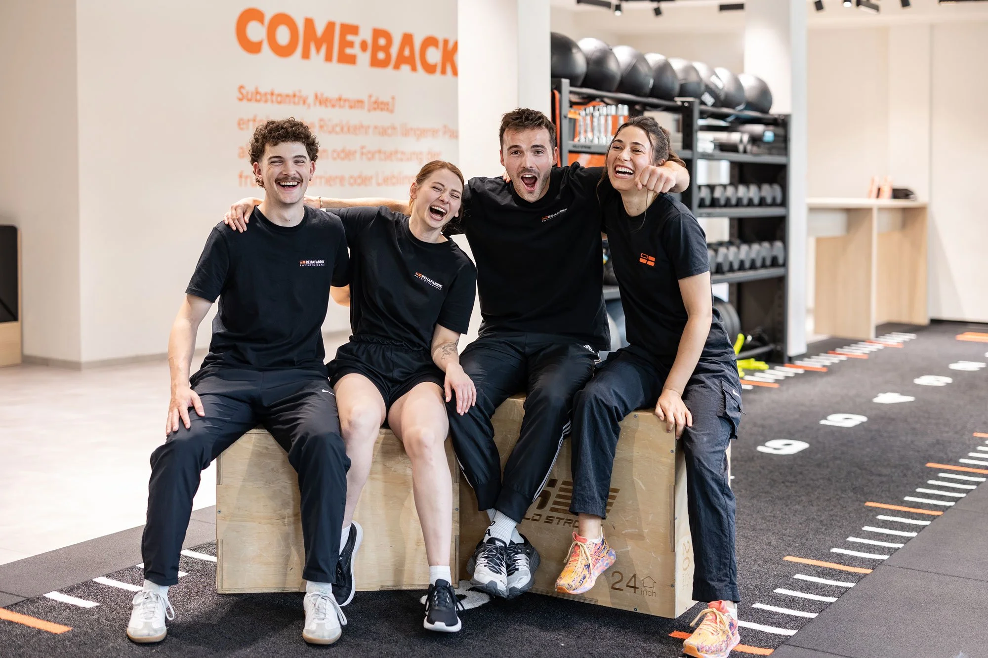Vier Physiotherapeut:innen sitzen auf Kisten auf der Trainingsfläche, sie umarmen sich und lachen.