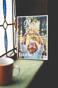 Das Mitglieder- und Mitarbeiter:innen Magazin  der AWO Weser-Ems steht auf der Fensterbank eines Buntglasfensters. Davor steht ein Becher. Auf dem Magazin steht der Schriftzug Marie und darunter ist eien Frau die mit der Hand ein Herz formt.