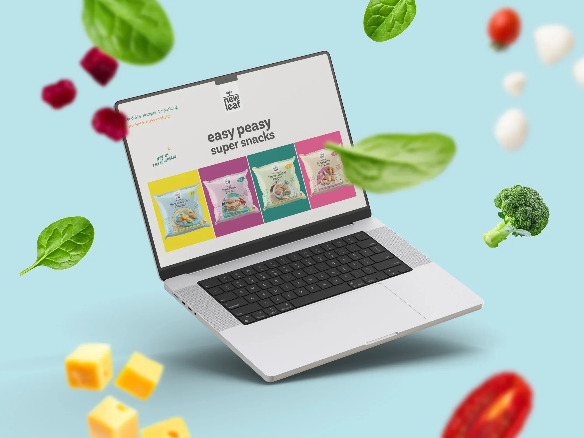 Ein Laptop zeigt eine Webseite mit dem Titel "easy peasy super snacks" und Produktbilder von Snack-Tüten. Im Hintergrund sind fliegende frische Gemüse- und Obststücke wie Brokkoli, Erdbeeren, Melanzani und Salatblätter auf einem hellblauen Hintergrund.