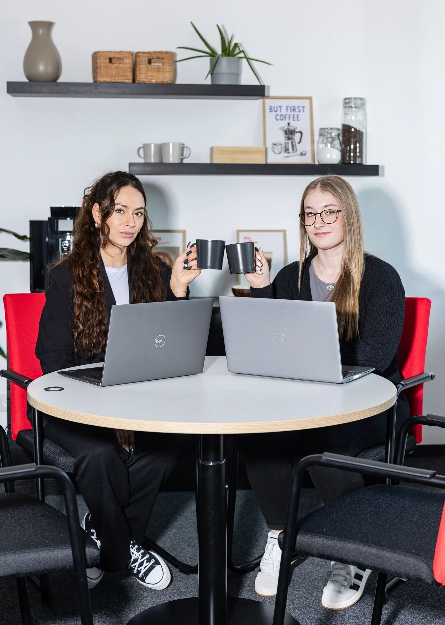 Zwei junge Frauen sitzen an einem Tisch in einem Büro, vor ihnen stehen Laptops und sie stoßen mit ihren Kaffeetassen an.