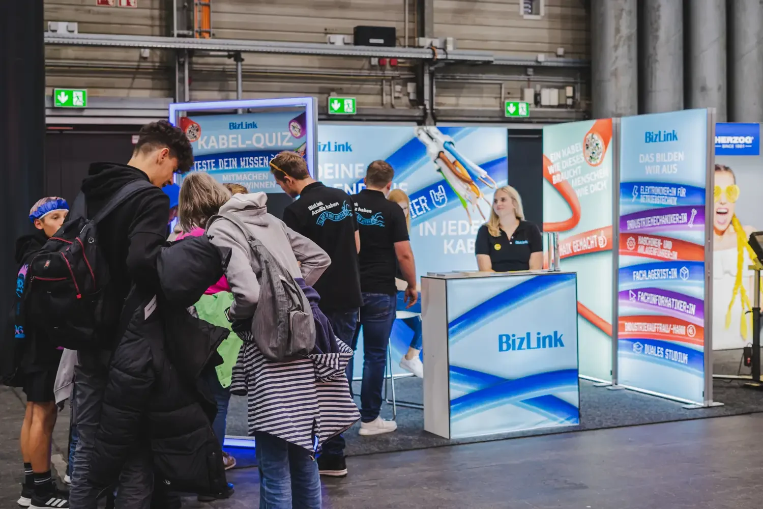 Menschen, die an einem Stand mit dem Logo BizLink auf einer Messe oder Veranstaltung teilnehmen, mit einem Hintergrund, der Werbebanner für Kabel-Quiz, Internet und Elektronik zeigt.