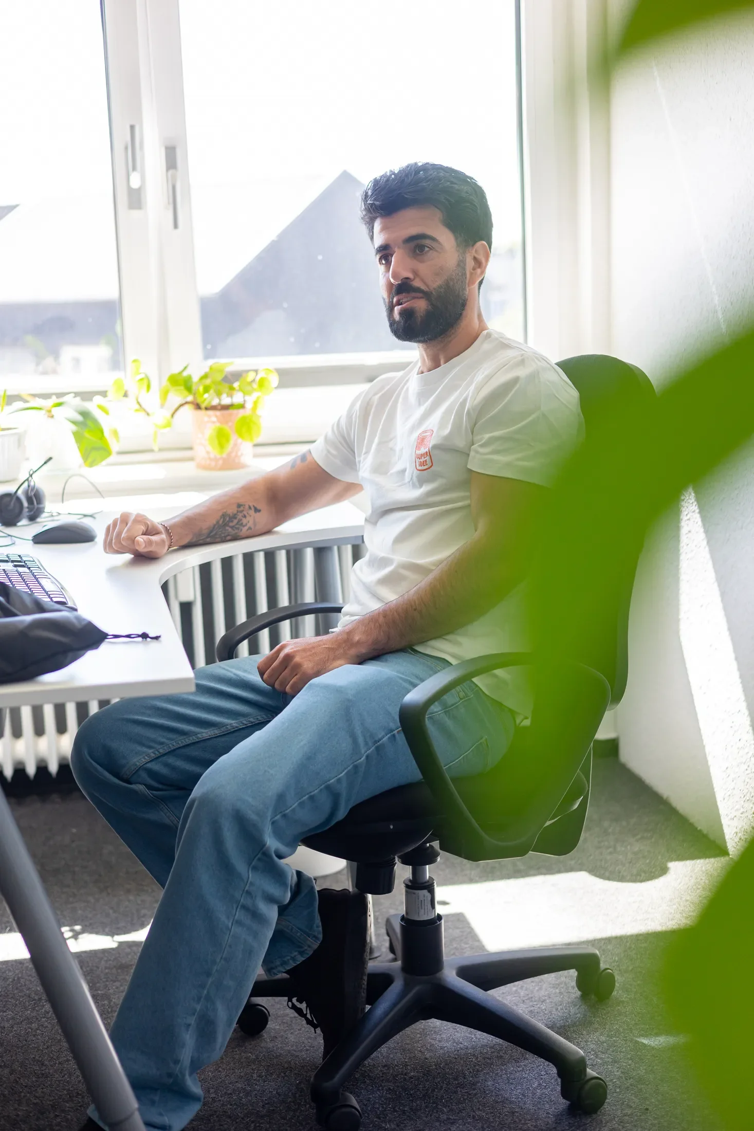 Ein Mann mit dunklem Haar und Bart sitzt in einem Büro, trägt ein weißes T-Shirt und blaue Jeans, vor einem Schreibtisch mit Pflanzen und Computertechnik, bei Tageslicht durch ein Fenster, grüner Blätter im Vordergrund.