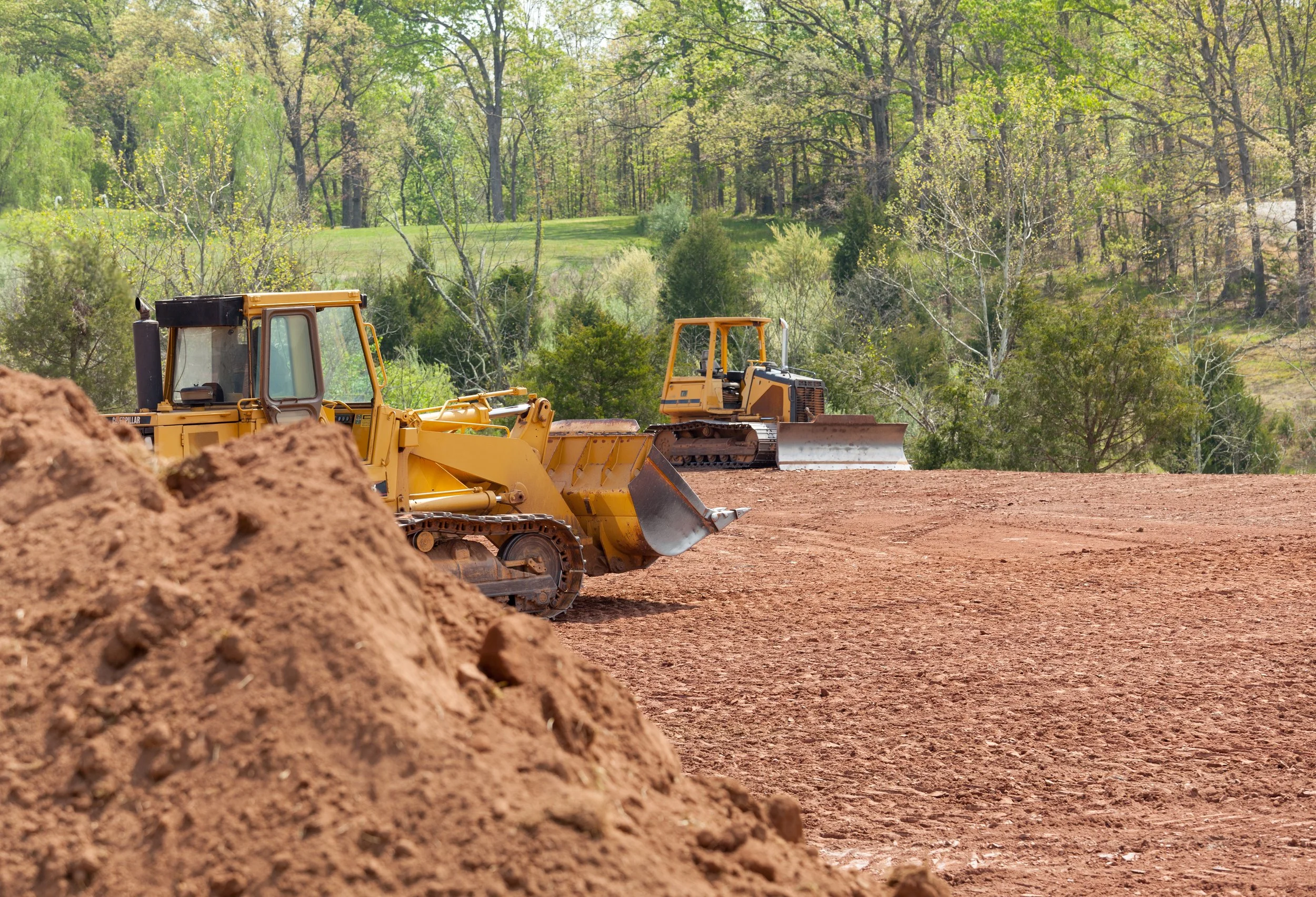 Two bulldozers working on a construction site with dirt in foreground and green trees and rolling hills in background.