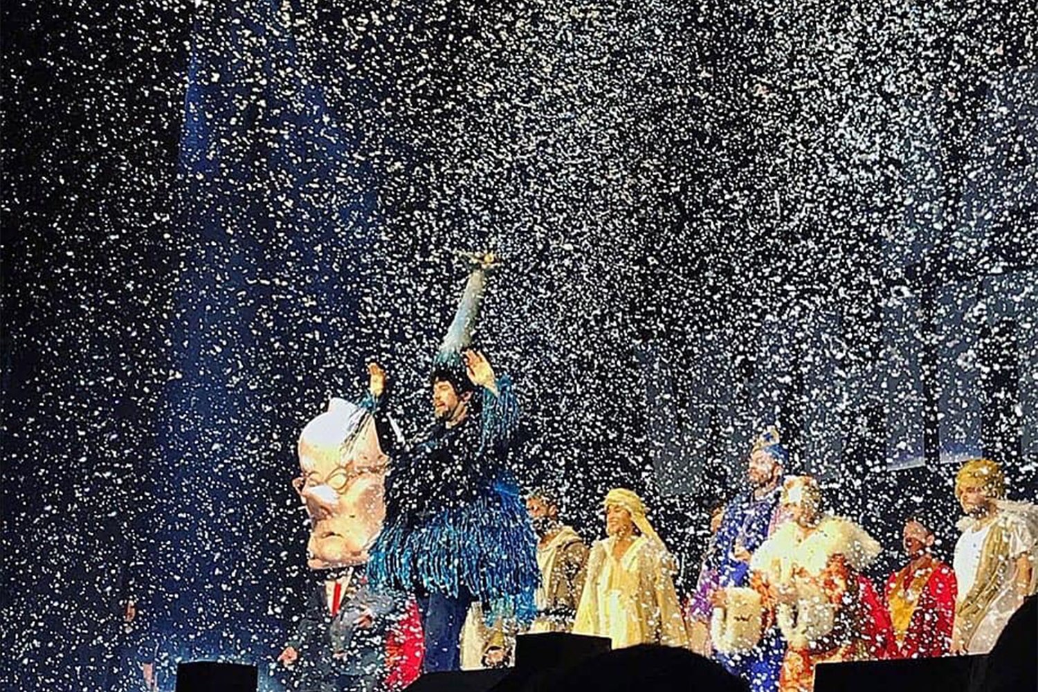 A stage performance with a man holding a puppet head and surrounded by performers in colorful costumes, with white confetti falling from above.