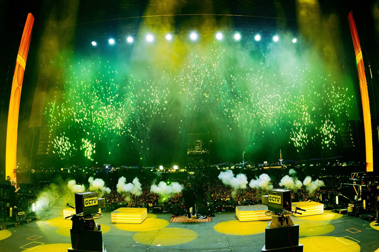Stage with bright yellow and green fireworks, smoke, and a crowd of spectators at night.