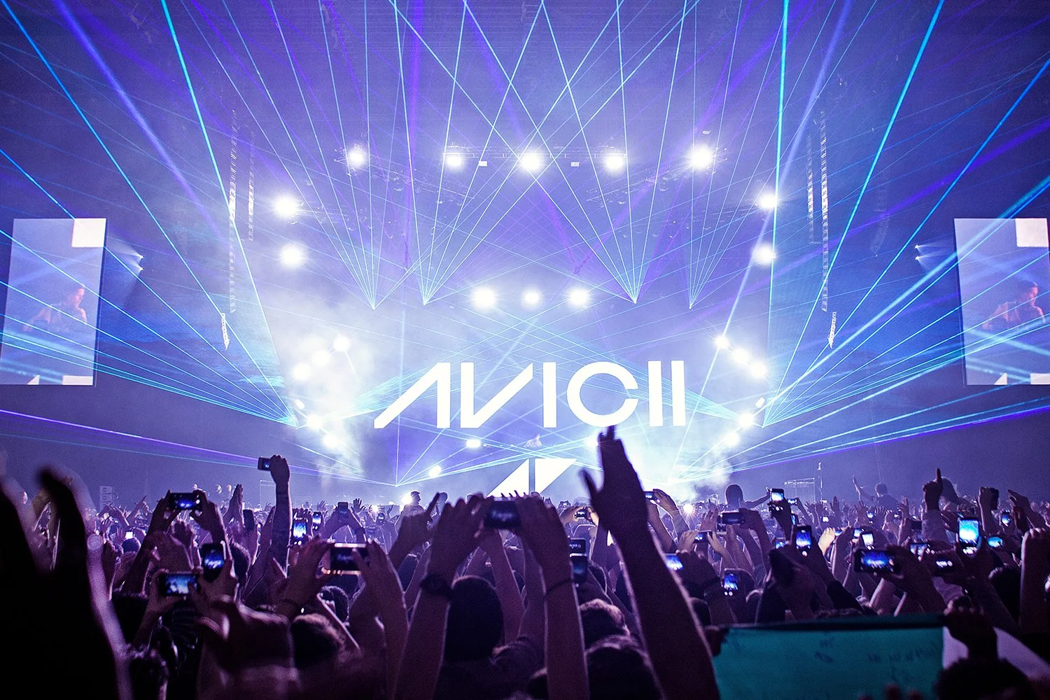 Crowd at a concert with lights and laser effects, featuring the text 'AVICII' on stage screens.