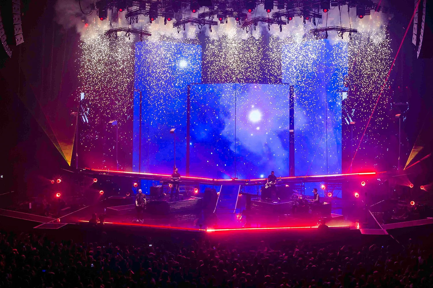 A concert stage with vibrant purple, blue, and red lighting, featuring digital screens displaying a galaxy scene and pyrotechnics creating sparks overhead, with a band performing in front of a large crowd.
