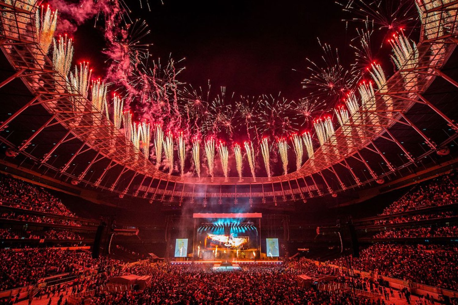 A large indoor concert venue with a crowd of people and fireworks lighting up the sky above the stage.
