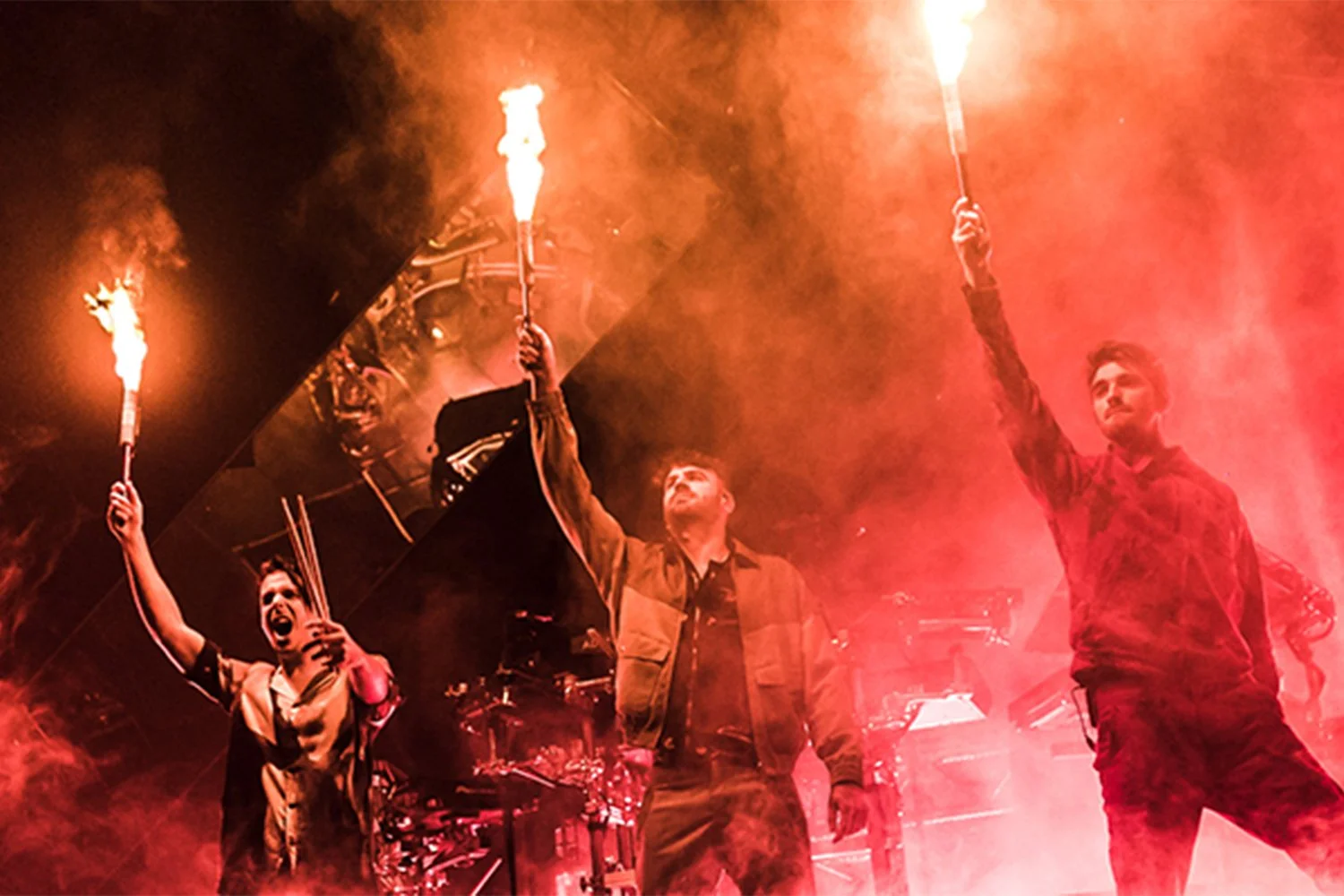 Three men on stage holding lit torches, with one man in the center and another on the right raising their torches upward, surrounded by red smoke and stage lighting.