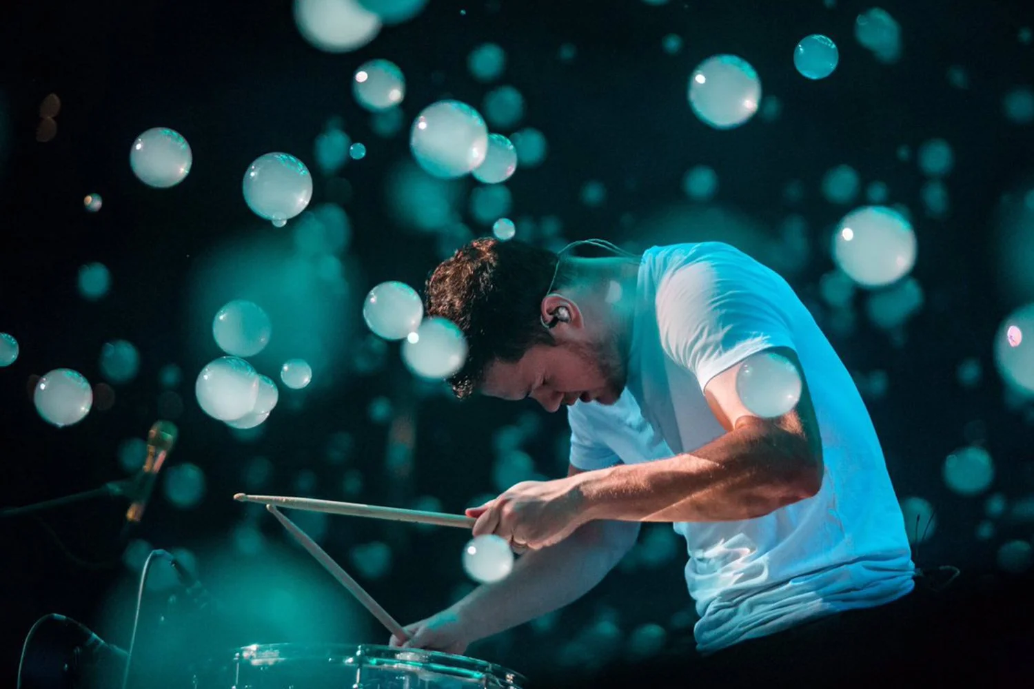 A man playing drums on stage with a dark background illuminated by floating orbs of light.