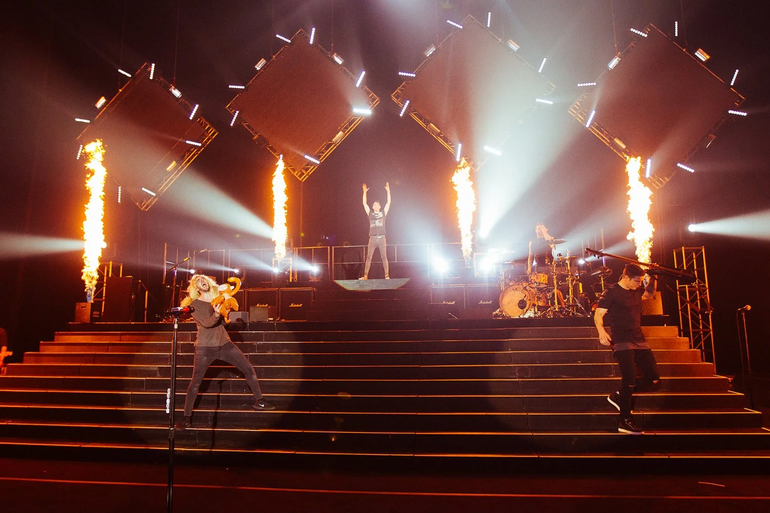 A live rock concert with a stage featuring bright lights, pyrotechnics, and three performers: a woman holding a plush toy and singing into a microphone, a man at the center raising his arms, and another man at the drums on the right.