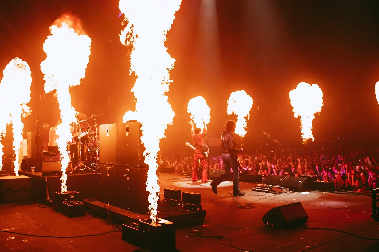 A concert stage with performers and pyrotechnics, showing fire bursts shooting upwards behind the band and crowd.