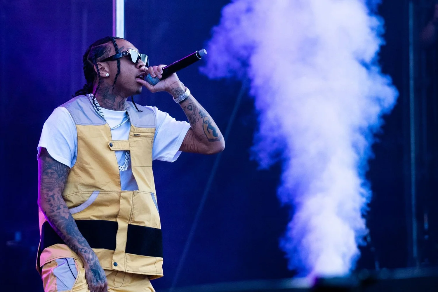 A man with tattoos, sunglasses, and jewelry singing into a microphone on stage with blue smoke in the background.