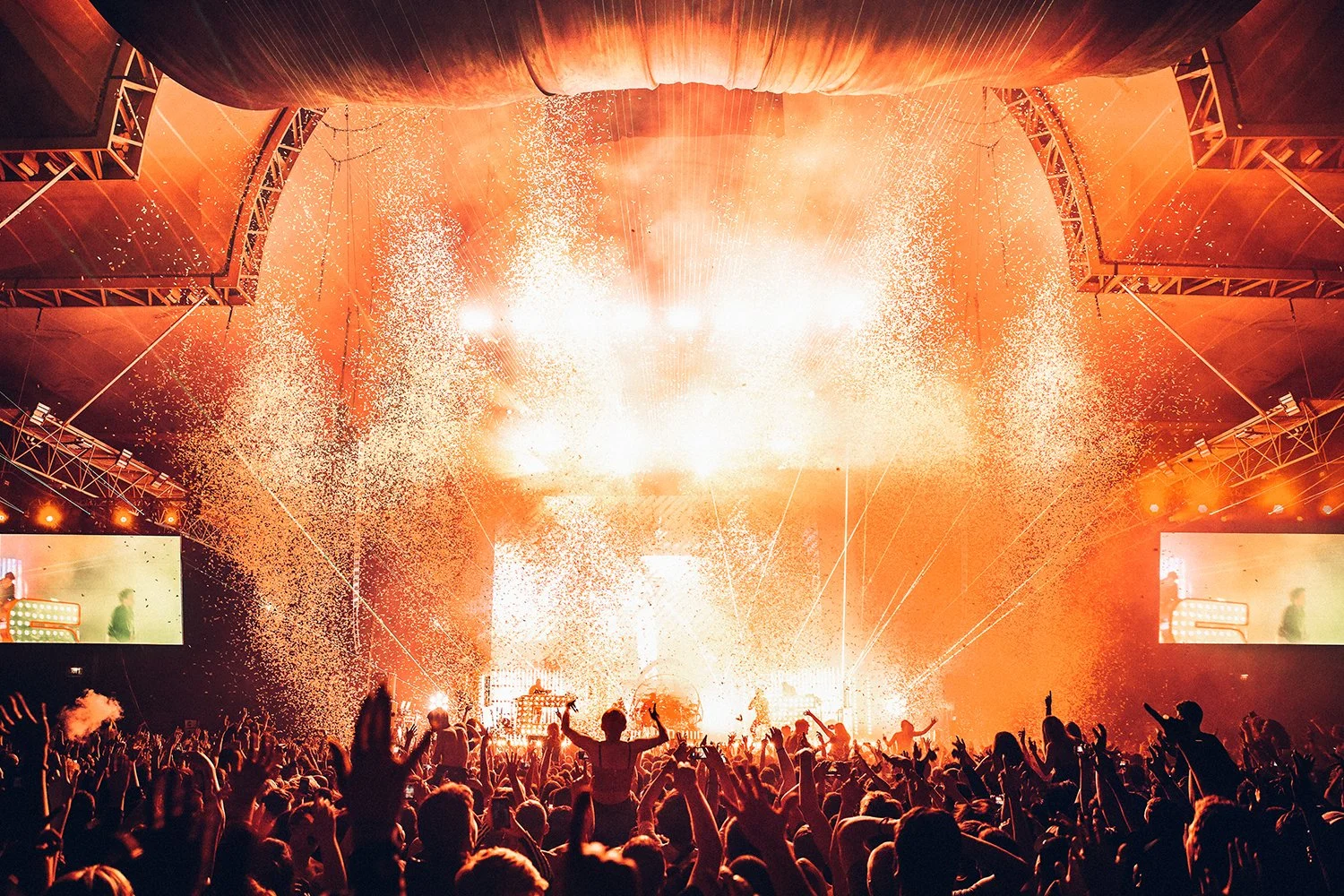 A lively concert with bright stage lights, pyrotechnics, and a large crowd of people dancing and cheering inside a venue with a high ceiling and large screens showing the performers.