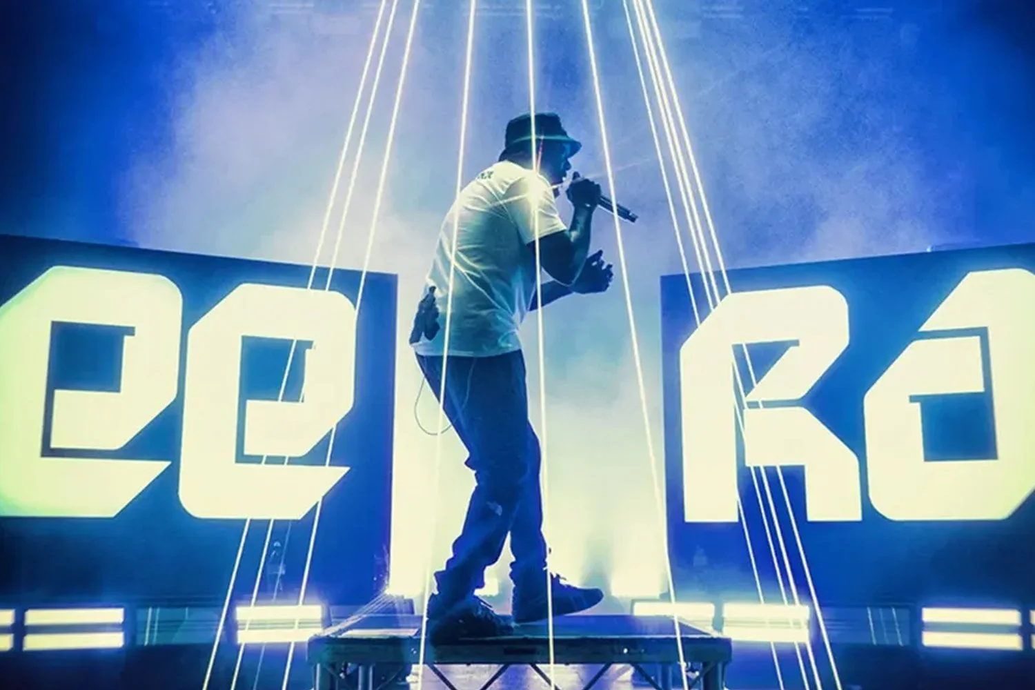 Performer on stage with large illuminated signs reading '20K' behind him, performing with a microphone in hand, surrounded by blue and white lights and laser effects.