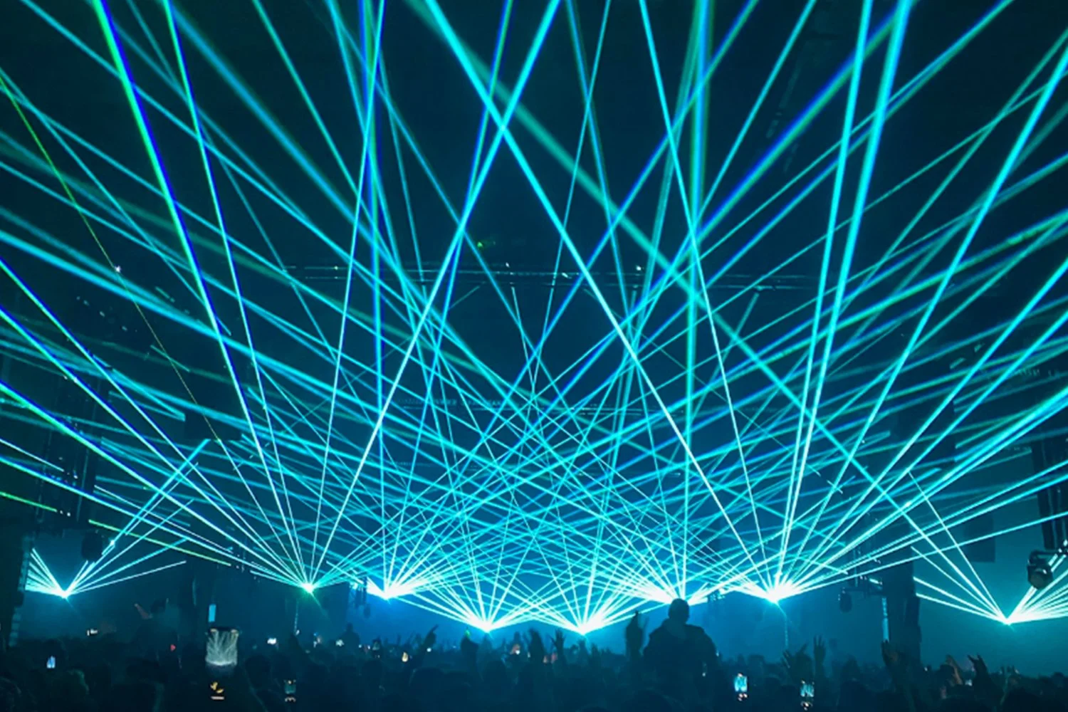 Blue laser lights crisscross over a crowd at a concert or event, with audience members raising hands and taking photos.